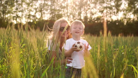 El-Concepto-De-Una-Familia-Feliz.-En-El-Campo-De-Centeno,-El-Niño-Camina-Por-El-Campo-Bajo-El-Sol-Poniente-Mirando-A-La-Cámara,-Mamá-Está-Detrás