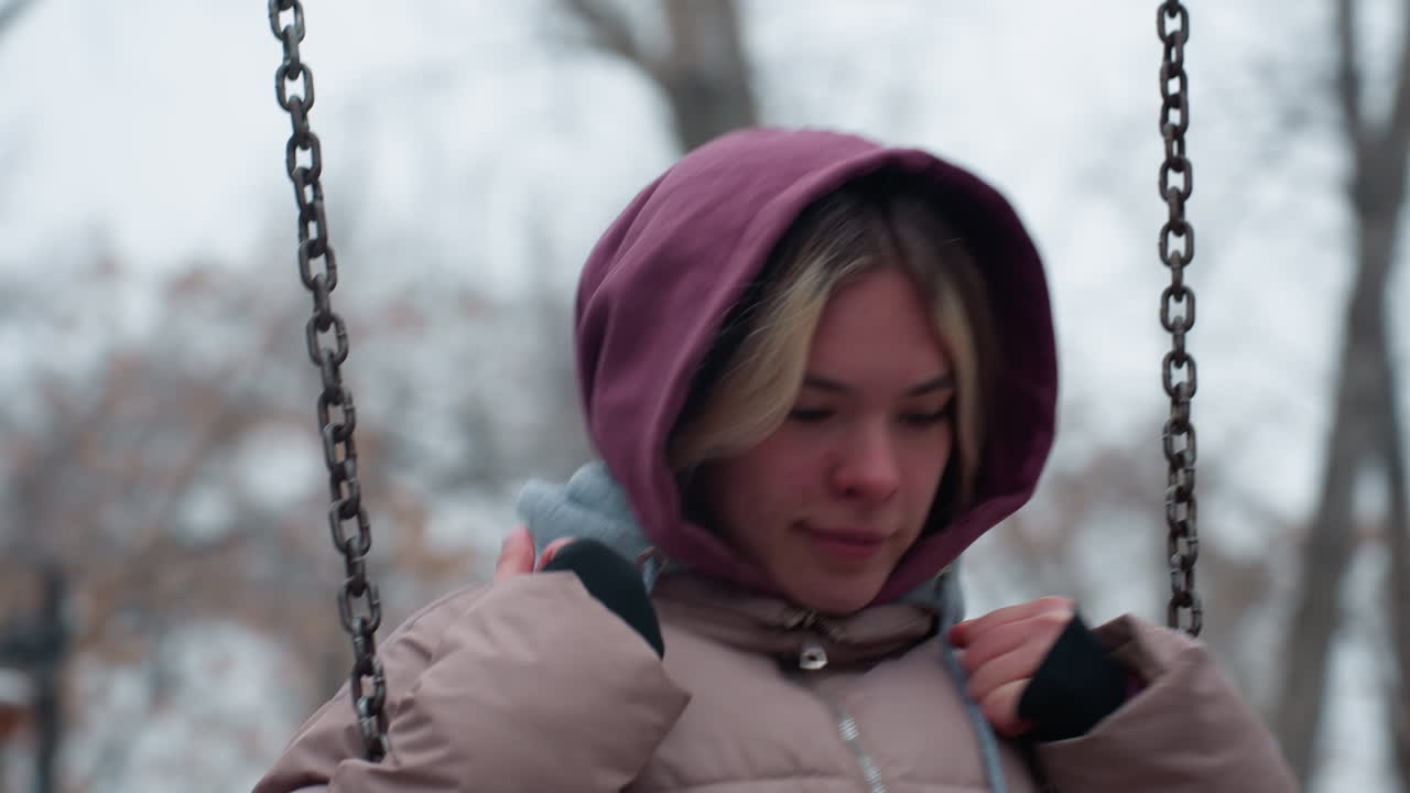 겨울 옷을 입은 젊은이가 스 체인을 들고 멀리서 사려 깊게 쳐다보고 있는 클로즈업, 배경에는 눈으로 인 흐릿한 나무와 건물이 있습니다.