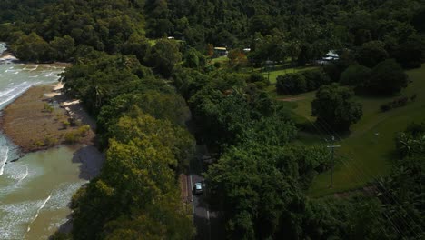 Aerial-drone-bird-view-of-modern-silver-car-driving-on-road-along-the-seaside-coast-with-sandy-beach-and-lush-green-tree-forest-and-blue-water-sea-in-4K