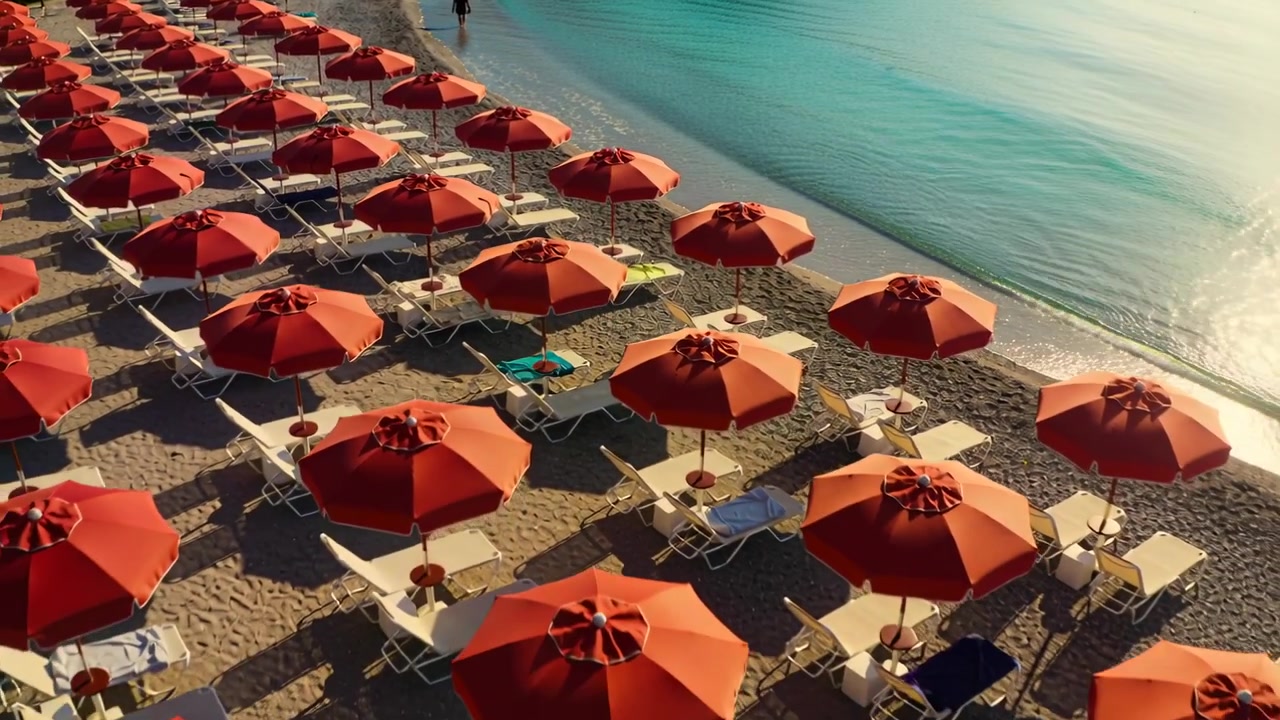 Un drone rivela file di ombrelloni arancioni lungo una spiaggia di ciottoli, seguendo una persona che cammina con abiti scuri.