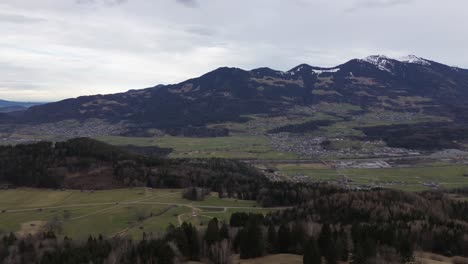 Drone-fly-above-forest-with-scenic-view-over-city-surrounded-by-snow-covered-summits-in-Vorarlberg,-Austria