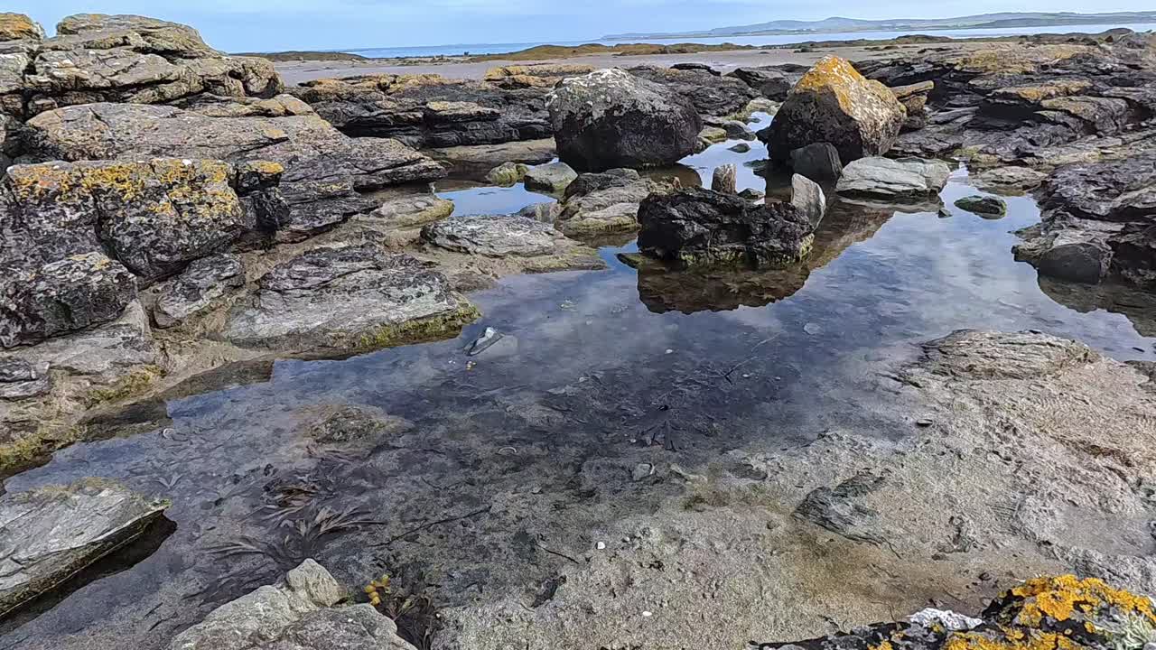 Shot of tidal pools in Penrhos Bay during low tide. Clear water reflects the blue sky while rocks and seaweed surround the scene