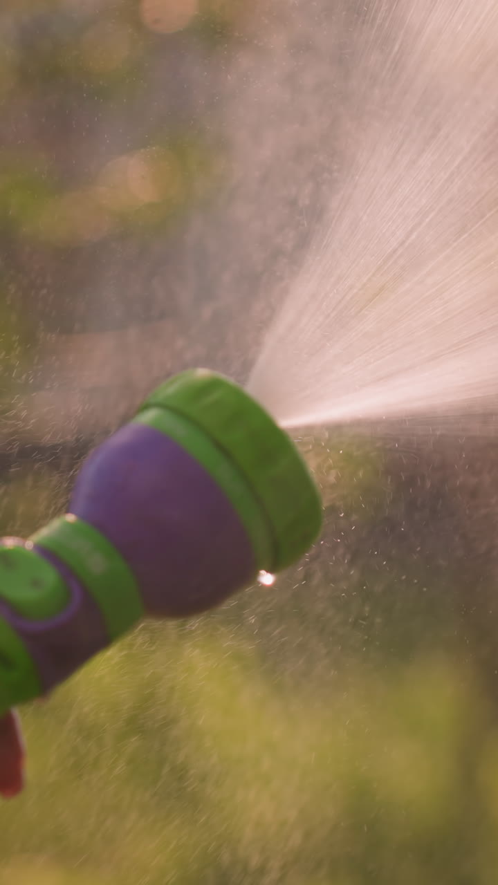 Close-up of hand holding water hose spraying fine mist outdoors, creating refreshing summer atmosphere, with sunlight reflecting on water droplets and soft-focused greenery in background