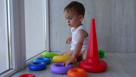 beautiful baby plays with toys sitting on the floor. kid is busy assembling a toy pyramid.