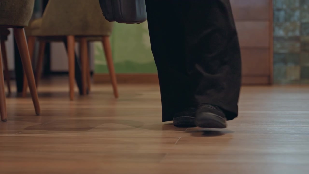 Closeup feet walking on wooden floor, black pants and dark shoes crossing cozy cafe interior with wooden chairs and warm lighting, person carrying briefcase, slow deliberate pace, polished parquet