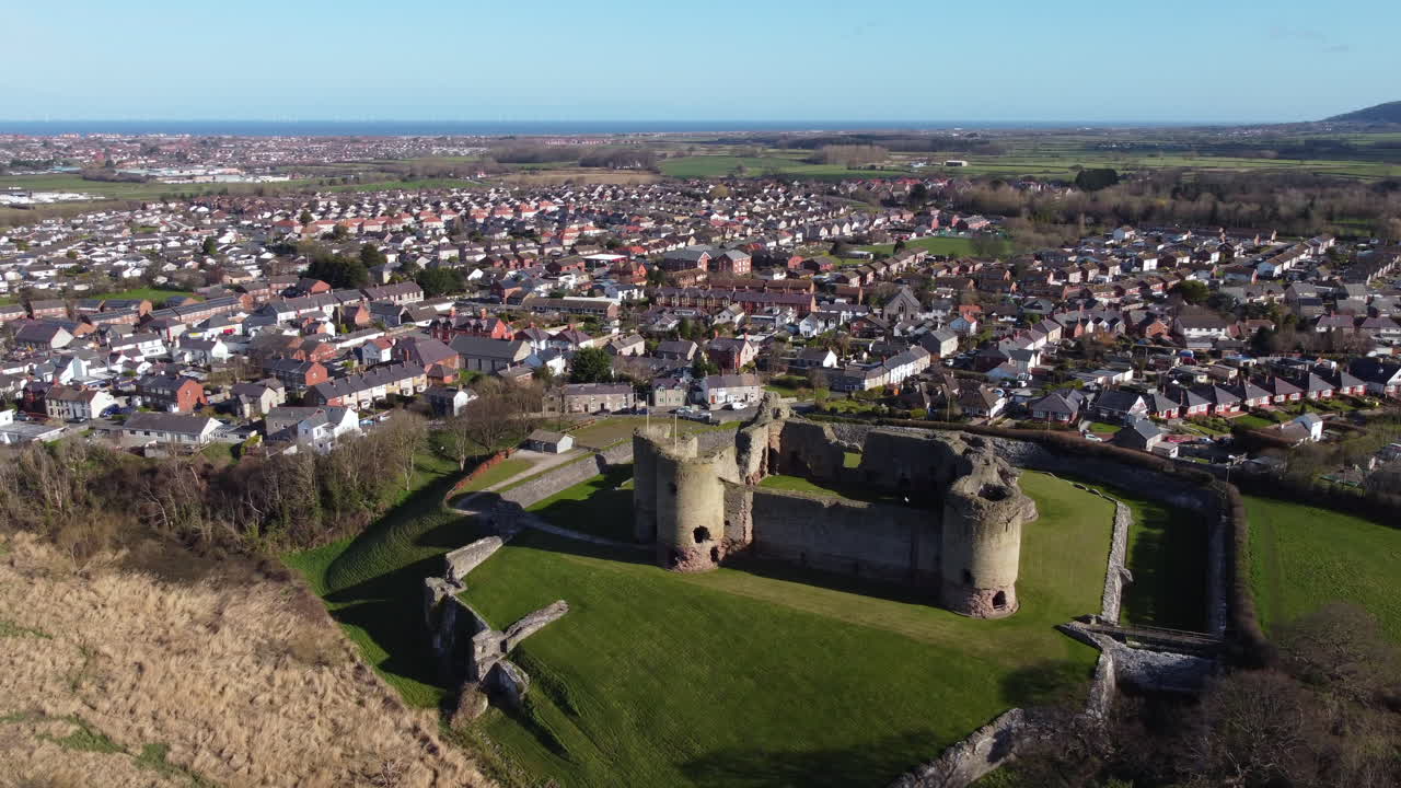 Una vista aerea del Castello di Rhuddlan in una soleggiata mattina di primavera, volando da sinistra a destra intorno al castello con la città di Rhuddlan sullo sfondo, Denbighshire, Galles, Regno Unito