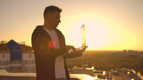 Mittlerer-Plan:-Ein-Männlicher-Freiberufler-Tippt-Bei-Sonnenuntergang-Auf-Der-Tastatur-Eines-Laptops-Und-Hat-Blick-Auf-Die-Stadt,-Während-Er-Auf-Dem-Dach-Eines-Wolkenkratzers-Steht.-Der-Geschäftsmann-Arbeitet-Aus-Der-Ferne-Und-Kontrolliert-Die-Arbeit-Des-Unternehmens