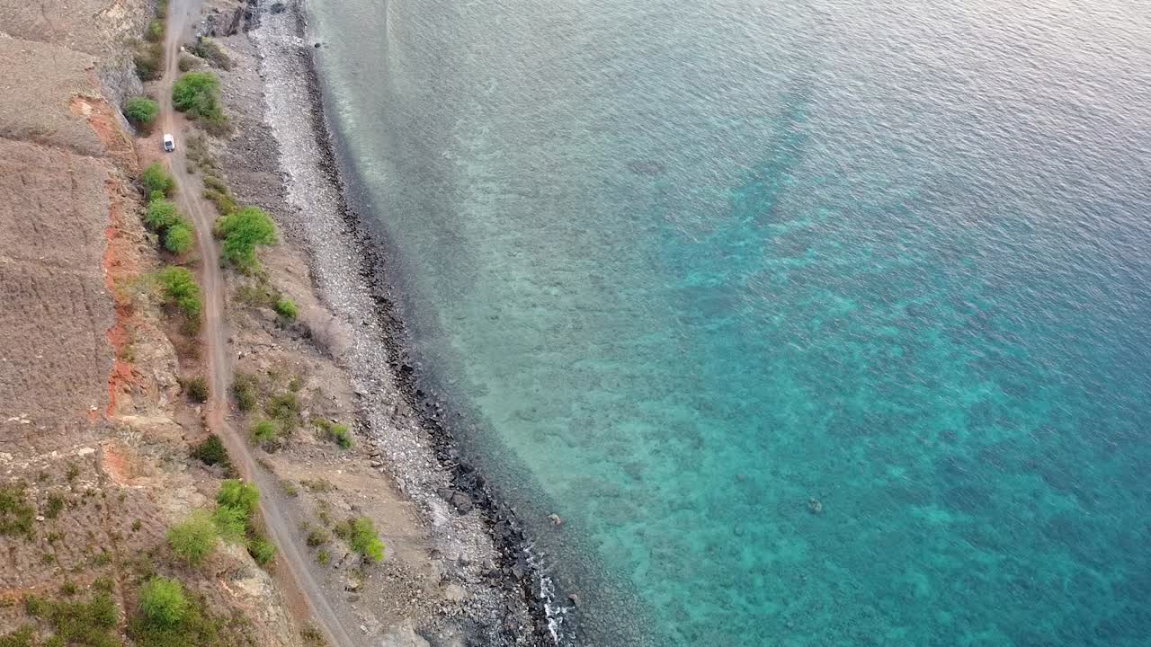 Aerial view of white car traveling along bumpy track on rugged coastal terrain with turquoise ocean water in Timor-Leste, Southeast Asia