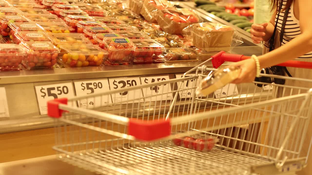 A person selects tomatoes in a well-lit supermarket aisle, placing them into a shopping cart