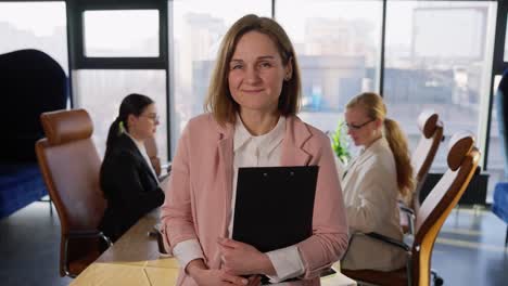 Portrait-of-a-happy-middle-aged-blonde-girl-in-a-pink-business-suit-with-a-tablet-in-her-hands-posing-near-the-table-in-a-modern-office-with-large-windows