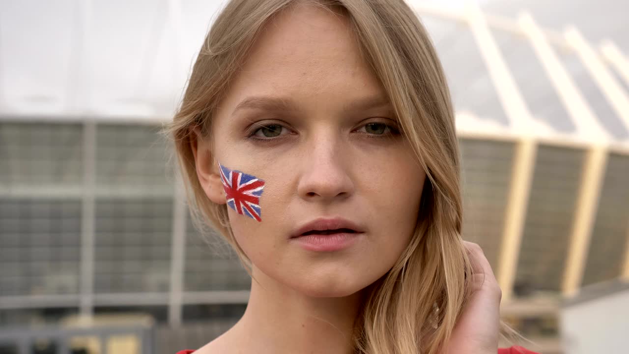 портрет молодой блондинки, футбольной фанаты из англии, смотрящей в камеру, серьезное лицо, стадион на заднем плане, 50 кадров в секунду.