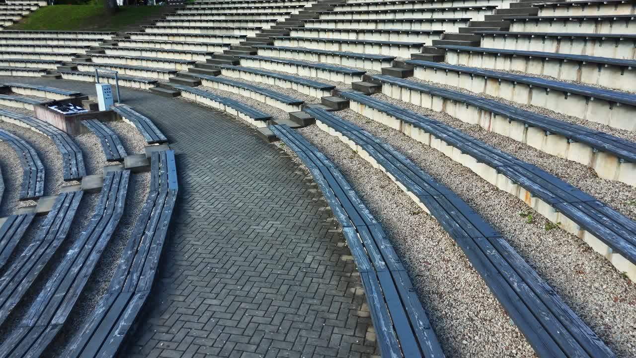 Weathered wooden benches curving along herringbone brick pathway, surrounding gravel ground in Sauleskalns amphitheater, forming peaceful outdoor seating area, drone pulling out