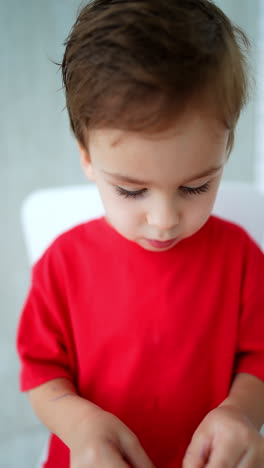 beautiful caucasian toddler boy in red t-shirt chewing food. kid peels an orange, shoves it into mouth and raises hands cheerfully. vertical video.