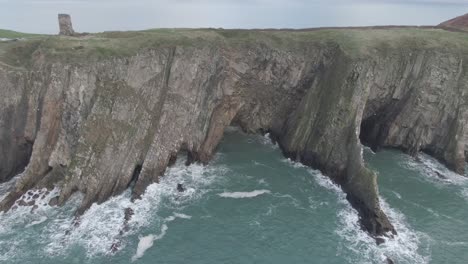 slow crane move up of rough sea side cliff with rough seas and waves hitting the cliffs reveling clear skies, green hills and castle ruin