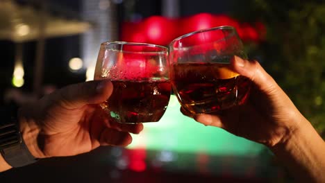 two people clink whiskey glasses in a celebratory toast beside a brightly lit outdoor pool at night, with warm, festive lighting and shallow depth of field