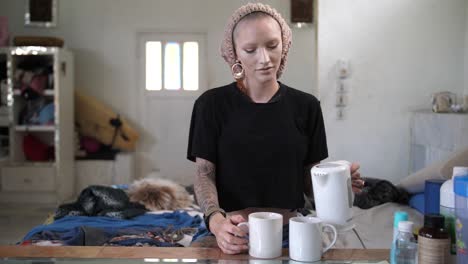 a young, white, alternative woman with a unique look in style and fashion puts two tea bags in two mugs and pours hot boiling water from the boiler into them, stands up and walks away