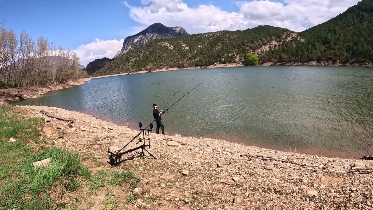 En fisker fisker aktivt fra den klippefyldte bred af en fredfyldt bjergsø, omgivet af frodige grønne bakker og fjerne blå himmel. Scenen indfanger naturens fredfyldte skønhed.