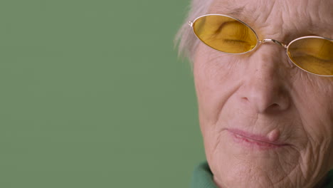 Close-Up-View-Of-Blonde-Senior-Woman-With-Short-Hair-Wearing-Green-Sweater-And-Sunglasses-Posing-On-Green-Background-Looking-At-Camera