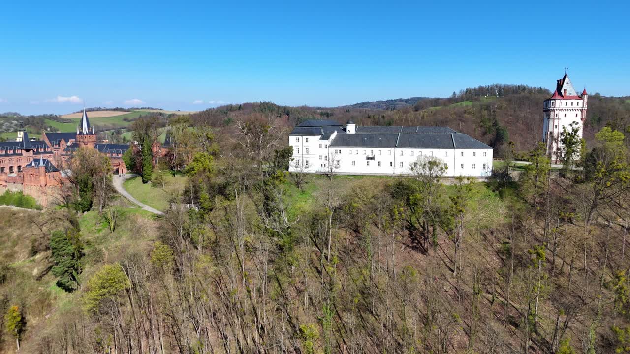 Volo fluido all'indietro mentre il drone ruota lentamente, rivelando il complesso del castello di Hradec nad Moravicí con edifici rossi e bianchi su una collina, circondato da alberi e campagna, sotto un cielo azzurro limpido.