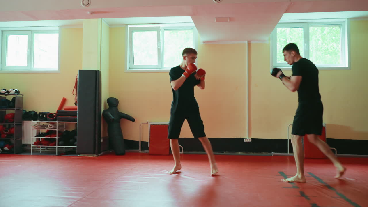 Two wrestlers training martial arts sparring in gym, facing each other on red mat, wearing gloves, barefoot, practicing combat stance, building strength, endurance, agility, under natural window light