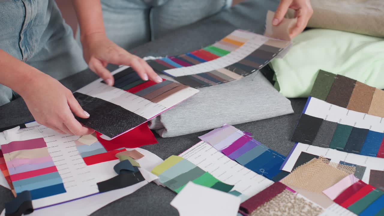 Elegant women selecting colors from vibrant fabric palette spread across work table filled with colorful textile samples, folded materials, paper patterns, and bright workspace background