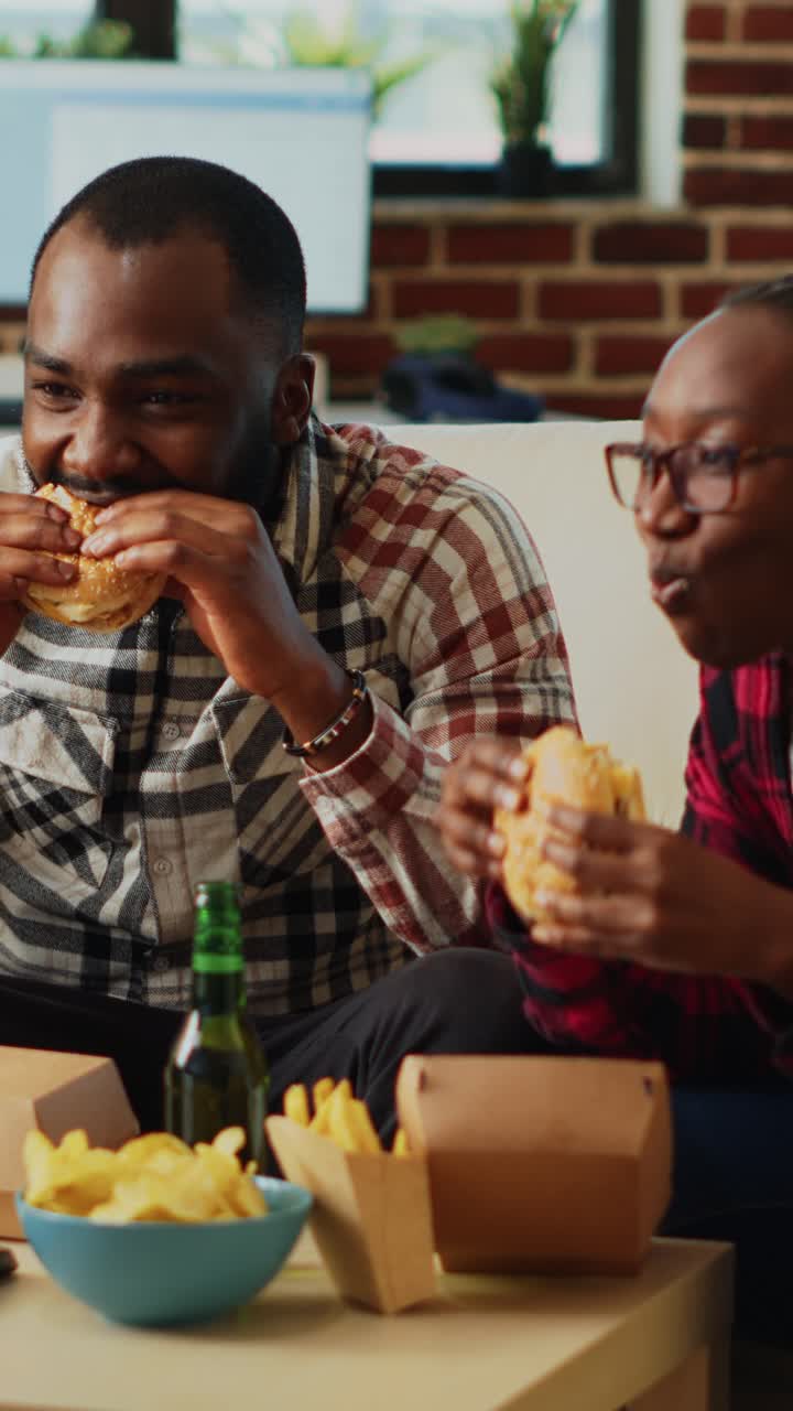 Video verticale: giovane coppia moderna mangia hamburger insieme sul divano