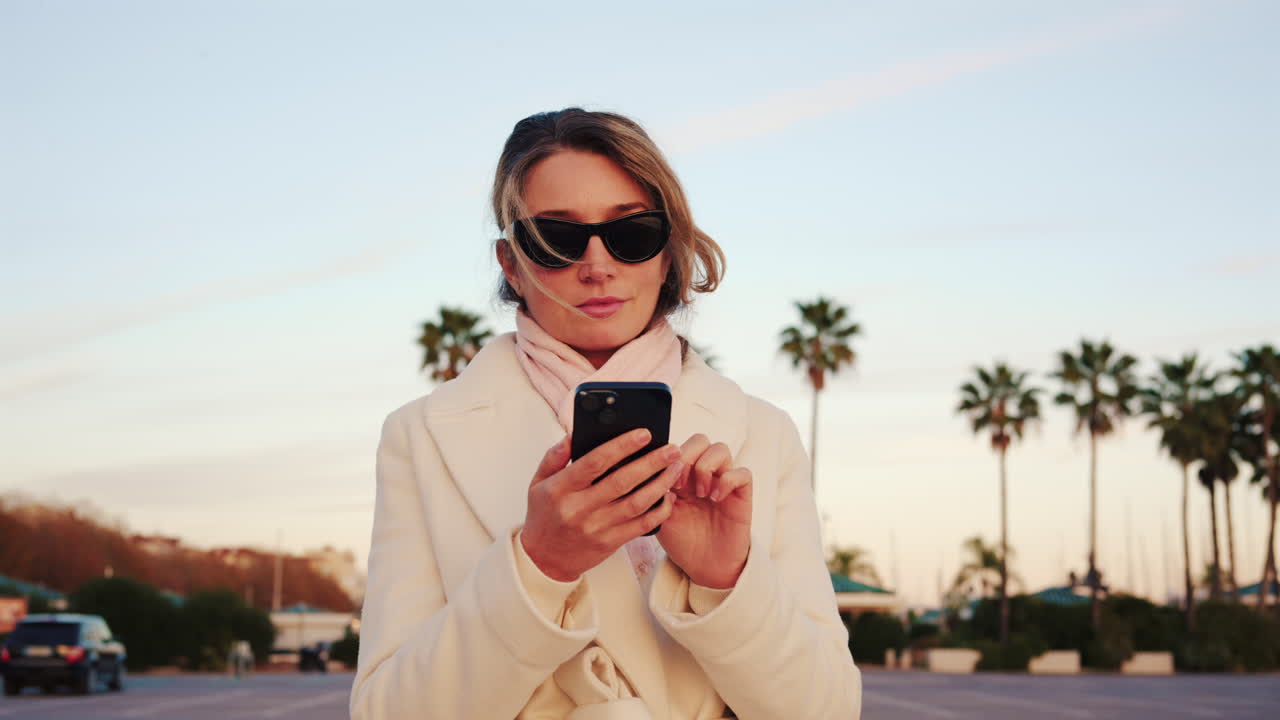 Donna elegante con cappotto bianco, sciarpa e occhiali da sole che controlla il telefono in una zona esterna soleggiata con palme