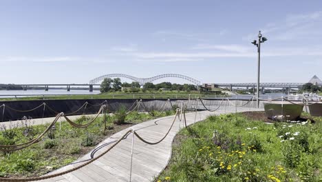 Camino-De-Madera-Rodeado-De-Plantas-Junto-A-Un-Río-Con-Un-Puente-Al-Fondo