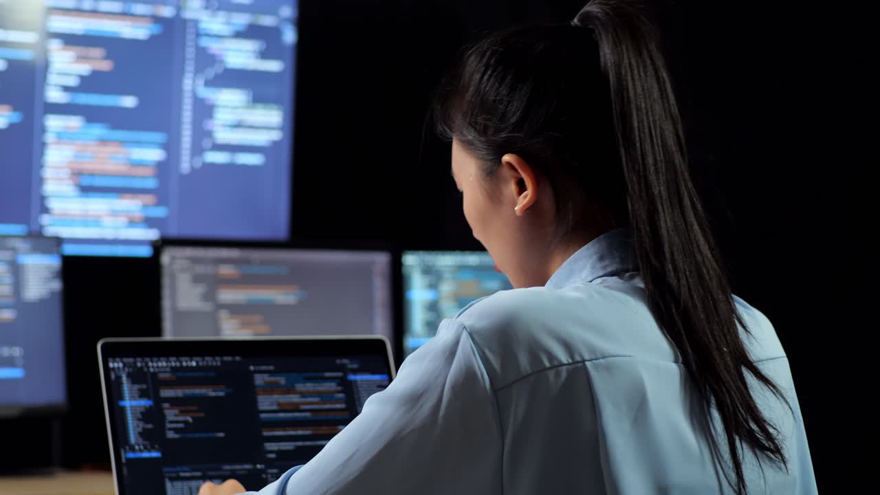 vista de cerca de una programadora asiática que tiene dolor de cabeza mientras escribe código en una computadora portátil usando múltiples monitores que muestran la base de datos en los escritorios de ventana de terminal en la oficina