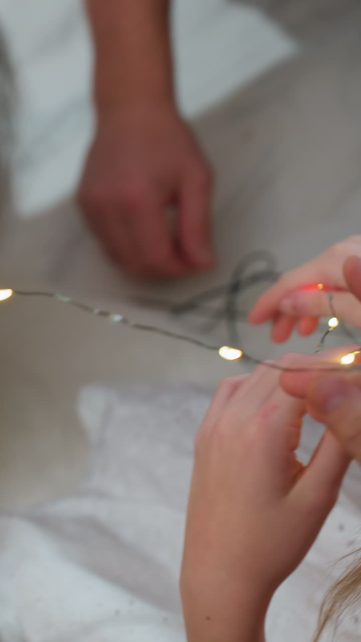 소녀가 머리핀 근처에서 빛나는 요정 전구 스풀을 들고 있고, 따뜻한 황금빛 전구가 달린 손과 머리 장신구를 클로즈업한 모습이에요. 아이는 장난스러운 장식을 위해 전구를 정리하고 있고, 보호자는 지켜보고 있으며, 부드러운 취침 시간 분위기예요.