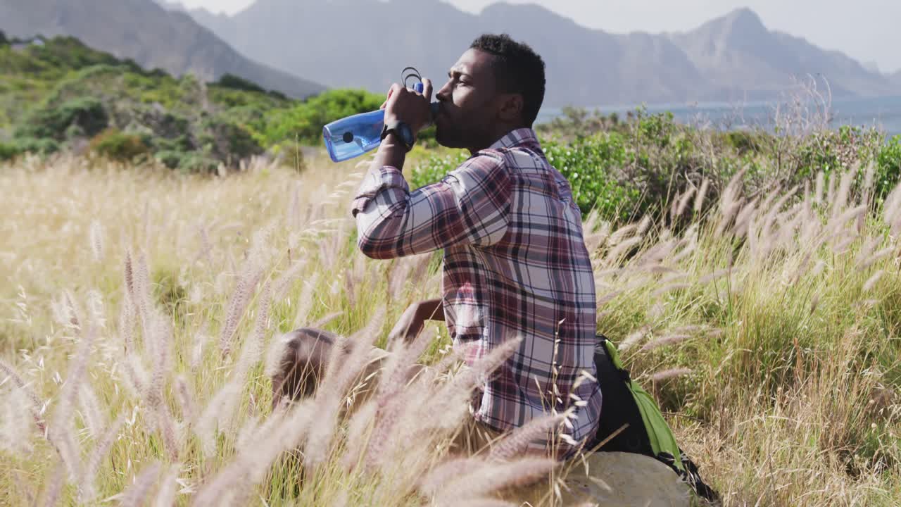hombre afroamericano bebiendo agua sentado en una roca mientras hace senderismo en las montañas