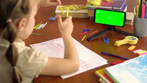 a preschool girl shows counting sticks, looks into a smartphone with a green screen. back view.