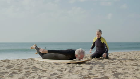 senior woman explaining to her husband how to surf while he lying on the surfboard on the sandy beach