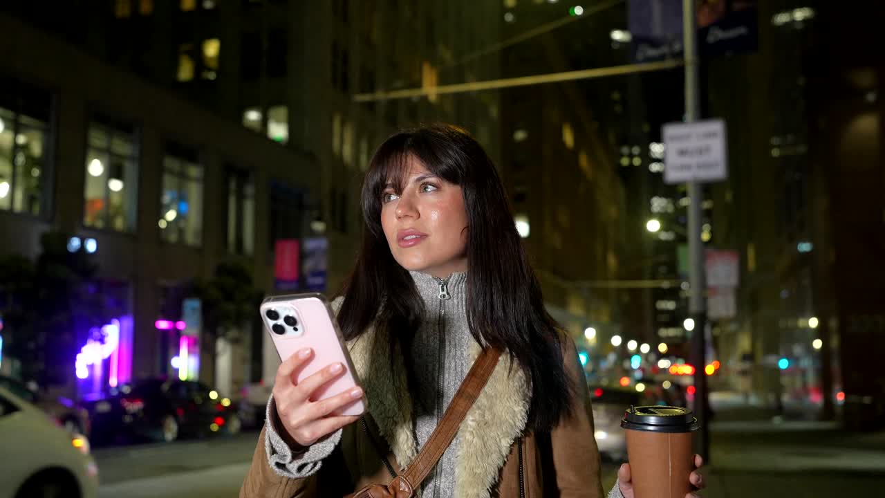 Giovane donna che usa lo smartphone e tiene in mano un caffè per strada di notte