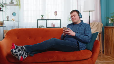 happy caucasian young man in wireless headphones listening music on smartphone lying on home couch