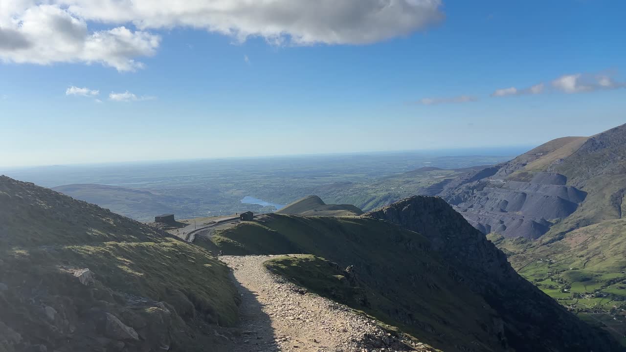 Mountain trail near Snowdon summit in Snowdonia shows steep cliffs, distant lake, and deep valley below under clear blue sky, capturing peaceful high-altitude view in early daylight