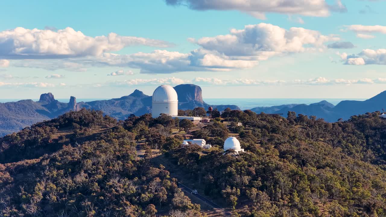 Drone footage glides over a forested mountain ridge with observatory domes, capturing clear skies, dramatic clouds, and distant peaks in warm evening light