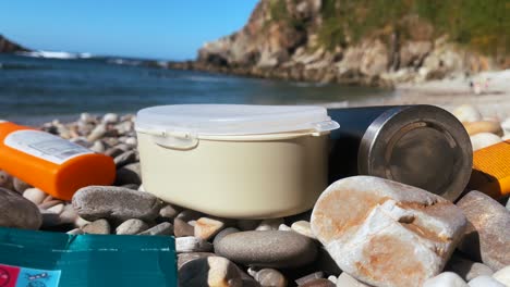 recipiente de plástico y botella de protector solar esparcidos en una playa de guijarros, contribuyendo a la contaminación