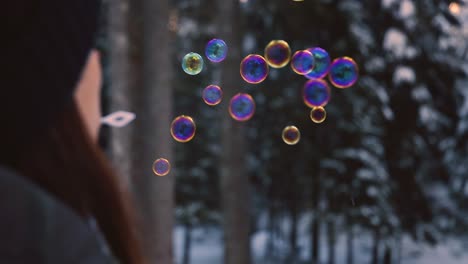 Brünette-Frau-Mit-Winterkleidung,-Die-Regenbogenfarbene-Seifenblasen-Bläst,-Die-In-Der-Luft-Schweben,-Mit-Einem-Verschneiten-Wald-Im-Hintergrund