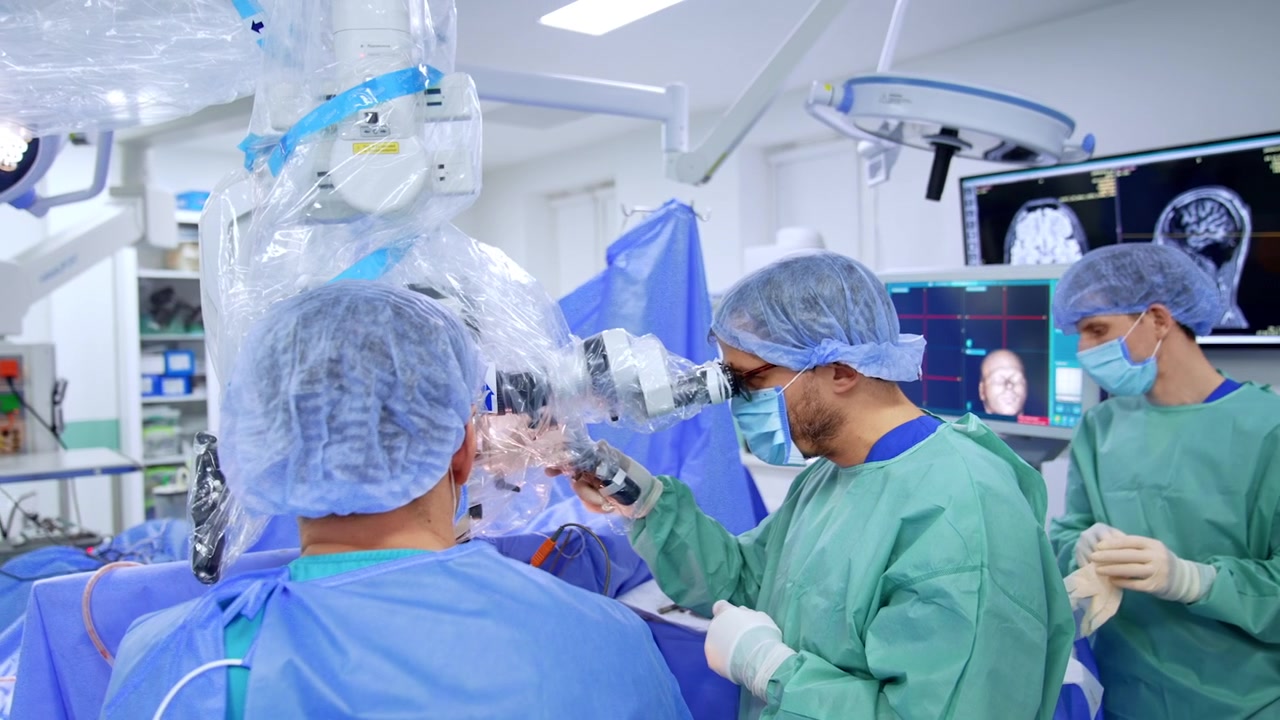 Neurosurgeon at microscope with assistant examining material during surgery. Surgeon in blue with loupe headband at eyepiece, teal gown assistant holds specimen at right.