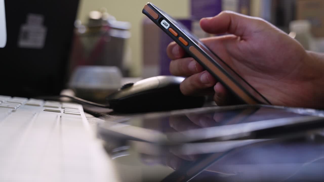 a hand holding a smartphone at an office desk beside a keyboard, mouse, and another phone, captured indoors with soft light and shallow depth of field