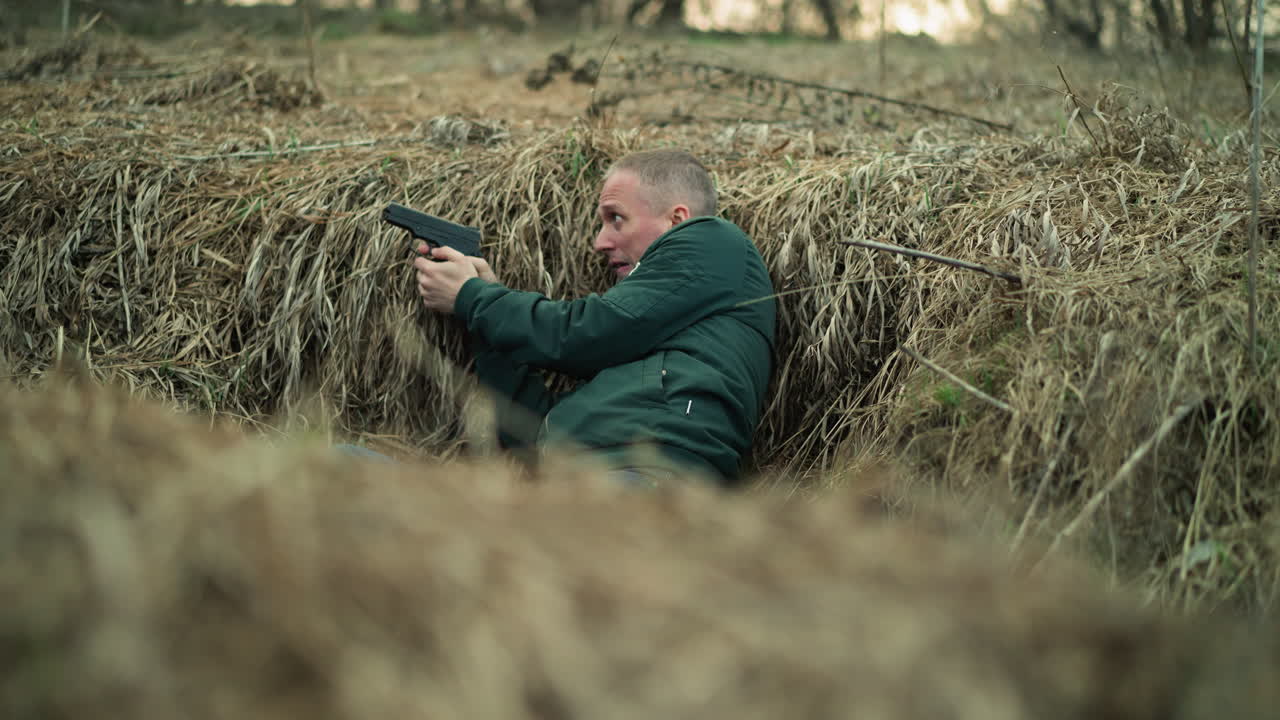 A close view of a man in a green jacket, lying in dry grass while holding a handgun and aiming with it and looking right