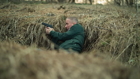 a close view of a man in a green jacket, lying in dry grass while holding a handgun and aiming with it and looking right