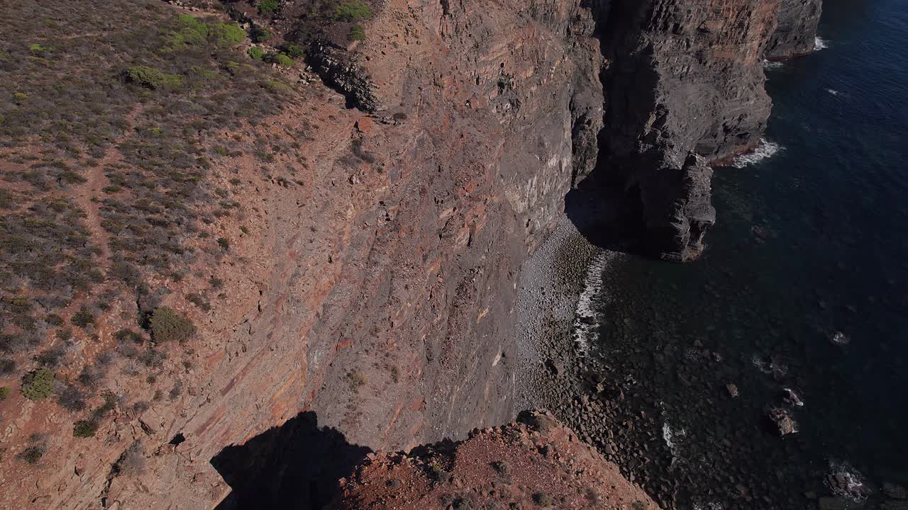 Scogliera sedimentaria atlantica a Vila Nova de Milfontes a Odemira, Alentejo, Portogallo, con parete rocciosa stratificata verticale che scende verso un litorale di ciottoli e acqua costiera limpida, ripresa con drone in discesa.