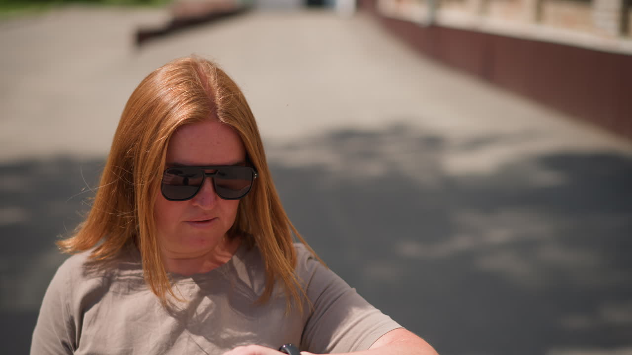 College lady with golden hair wearing sunglasses walking fast on sunny day checking time on smartwatch while looking around, soft shadows on pavement and blurred background