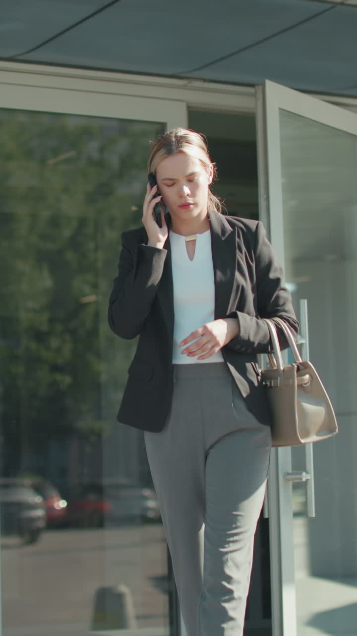 Lawyer exits mall on phone call with attentive expression, dressed in professional attire, holding handbag while walking, urban glass building reflects cityscape and vehicles