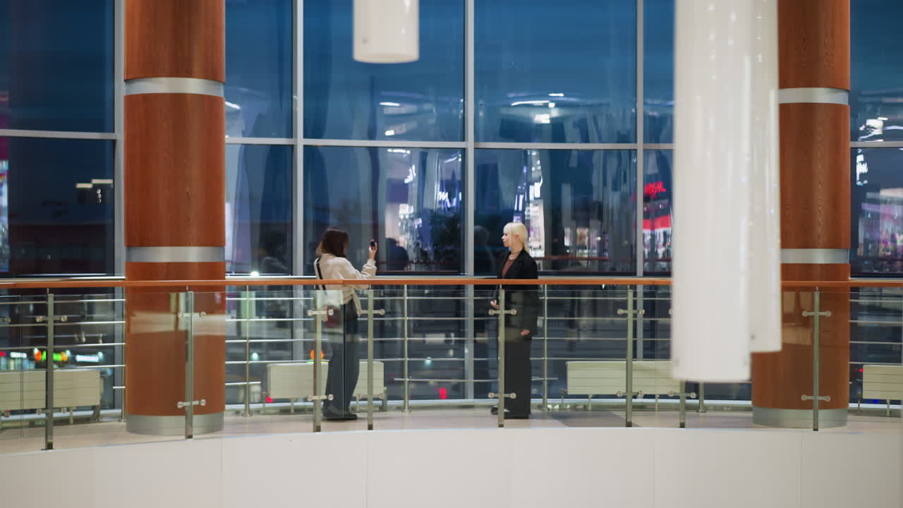 Two young friends standing indoors near large glass window with city lights outside as one holds phone capturing photo of other during evening in modern shopping center