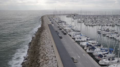 Convoy-dark-coloured-vehicles-travelling-along-wealthy-yacht-marina-harbour-aerial-follow-shot