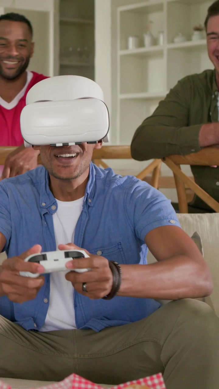 Video verticale: Giocatore VR in blu che si sistema le cuffie, tenendo un controller e ridendo con un amico in rosso a casa
