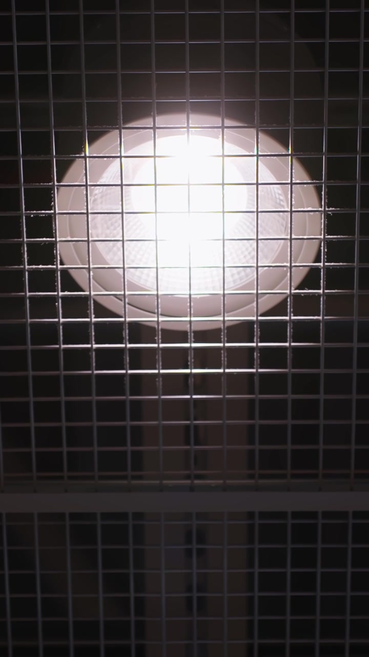 Pemandangan vertikal lampu langit-langit bandara yang bersinar menembus panel kisi logam, menyoroti garis geometris, infrastruktur dalam ruangan, dan desain terminal minimalis